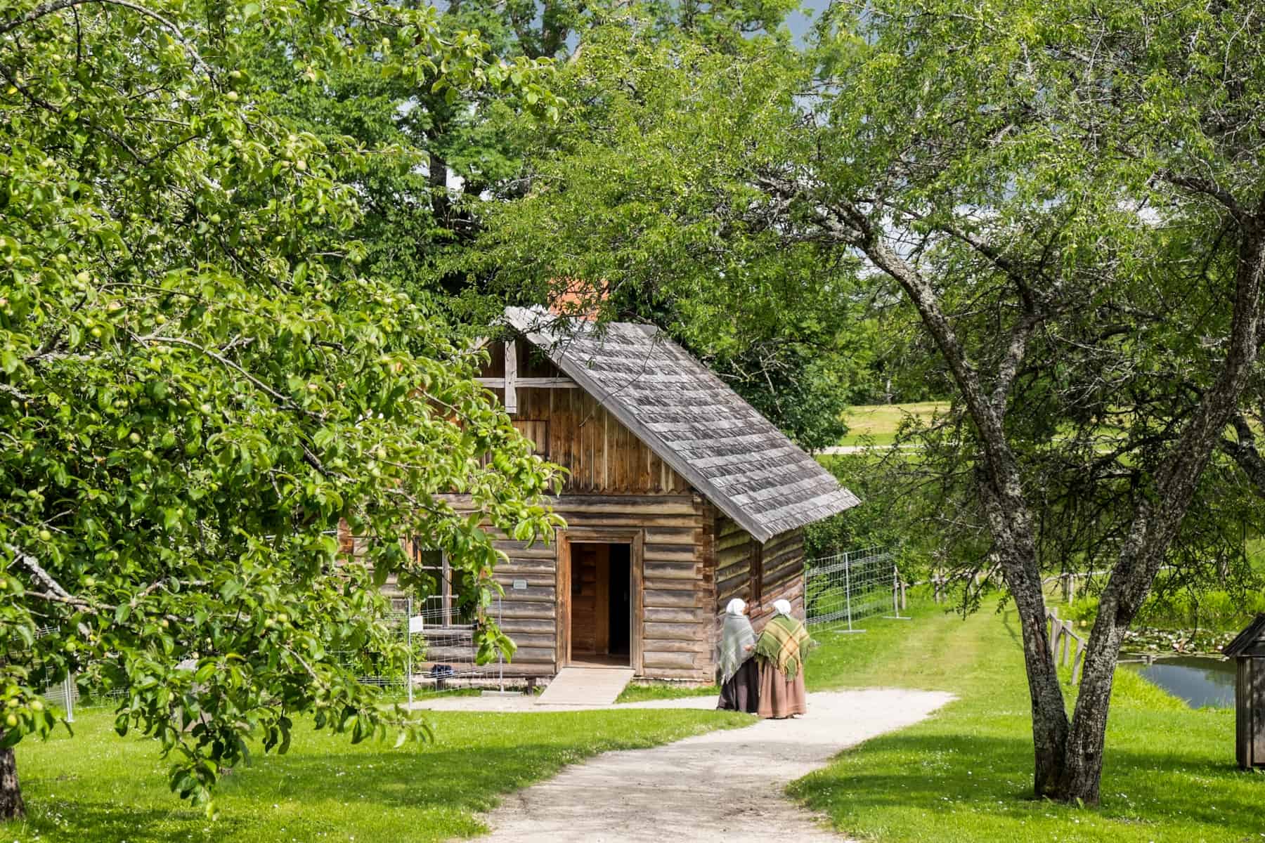 Two women dressed in traditional Latvian dress, with white headpieces, stand outside a wooden house with a triangular roof, within a field of trees.