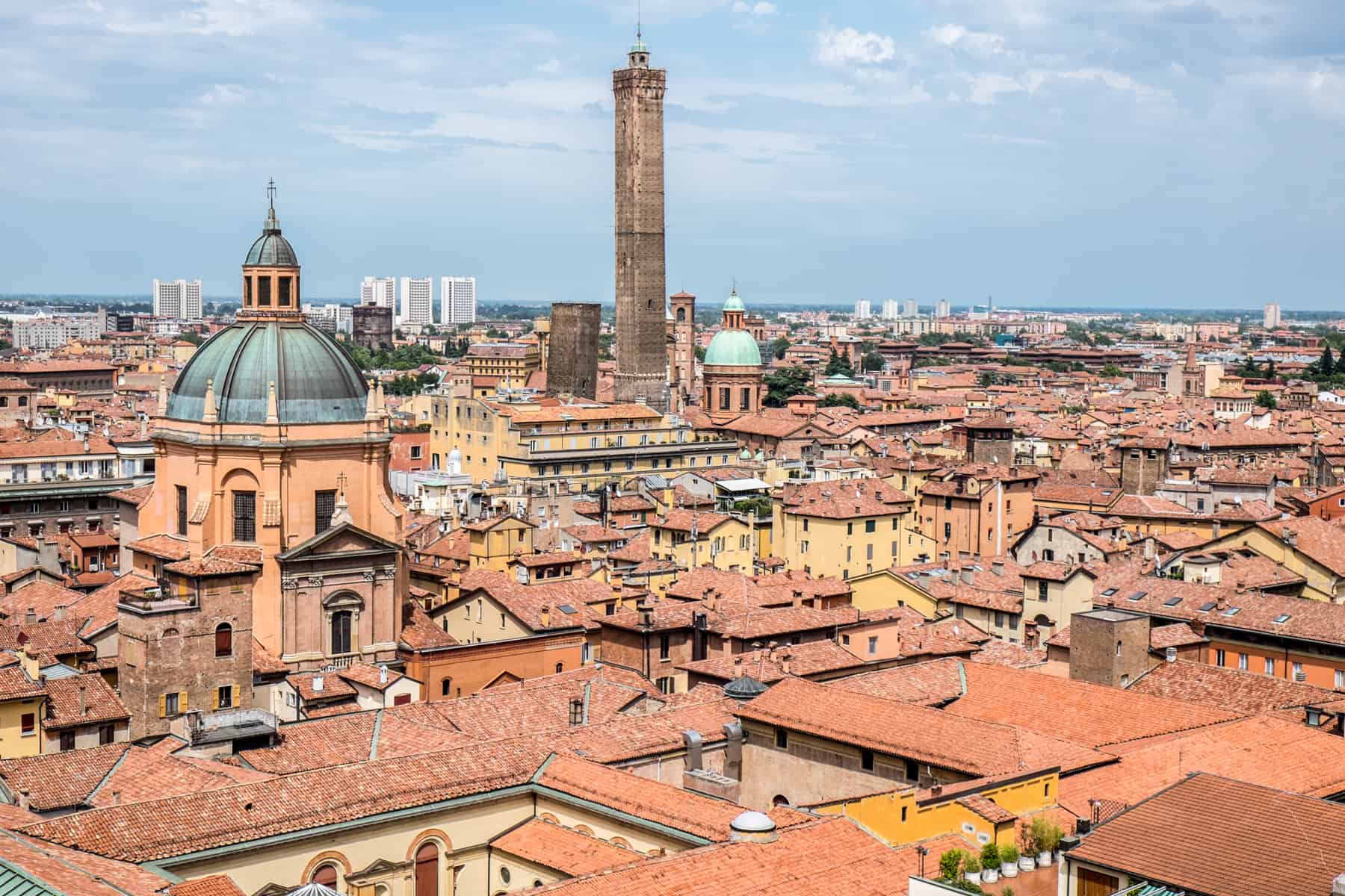 Elevated view of Bologna city over orange and yellow houses and the two stone towers that poke through. 