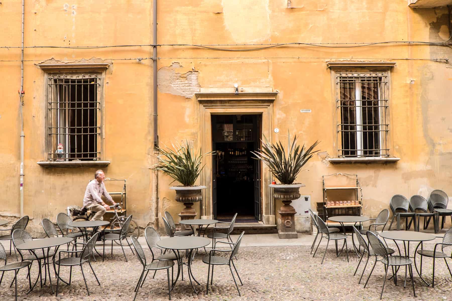 A man rides a bike past a rustic, yellow painted building with chairs and tables in front of it.