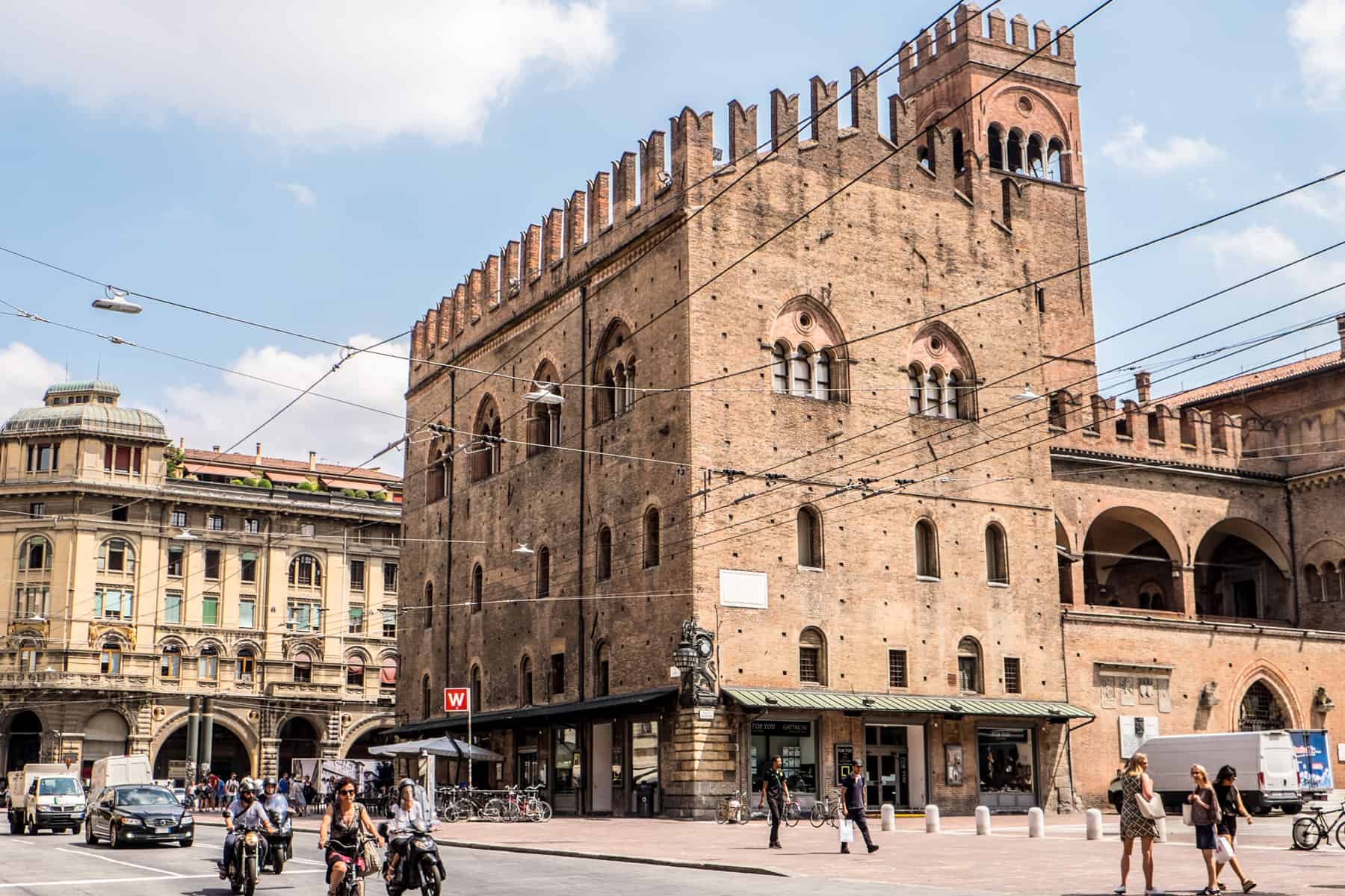 The brown stone, castle-like building next to a main road, is one of the main medieval structures in Piazza Nettuno in Bologna, Italy. 