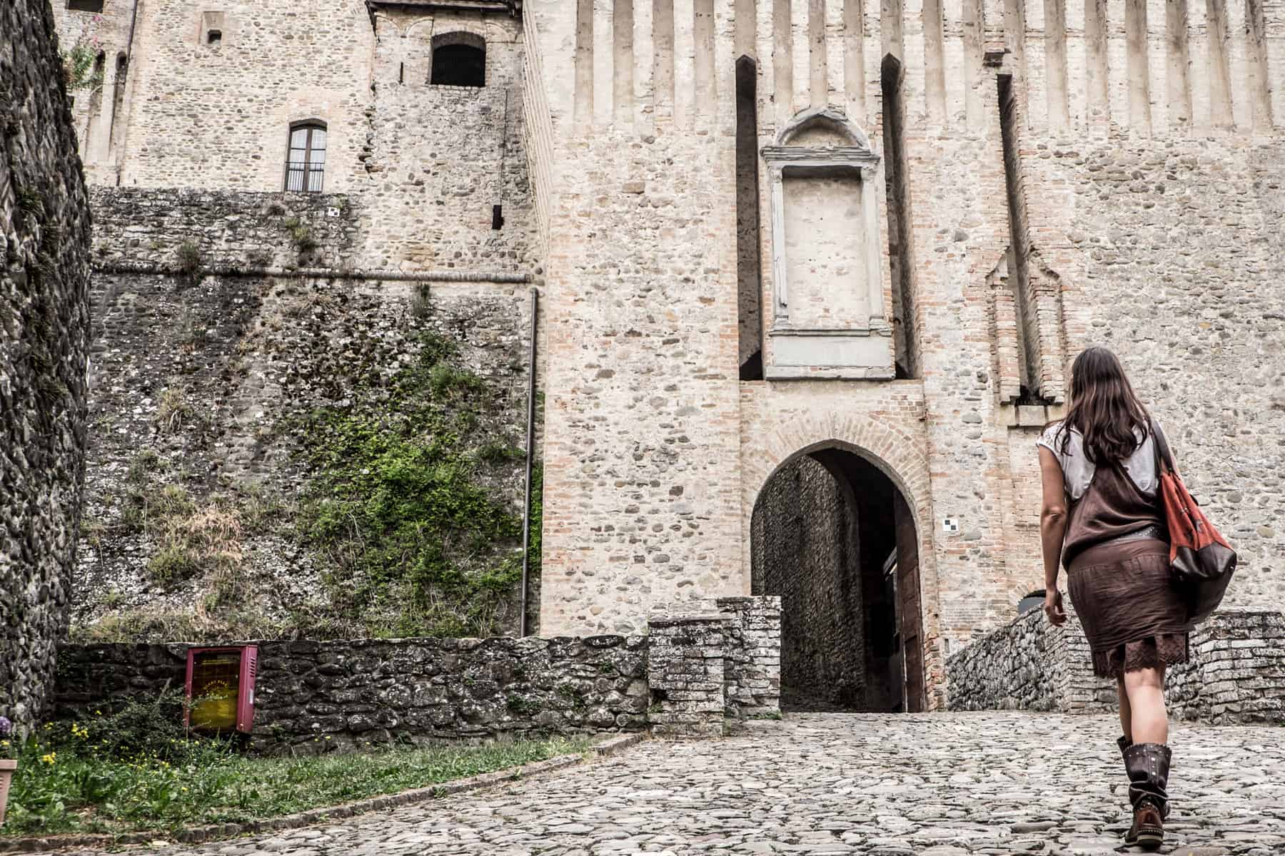 A woman in a brown dress and boots walks towards the arched doorway of a white and grey stone castle complex. 