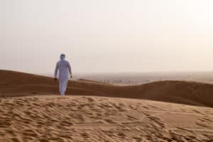 A man in a white robe walks through golden sand dunes in the Ras Al Khaimah desert at dusk.