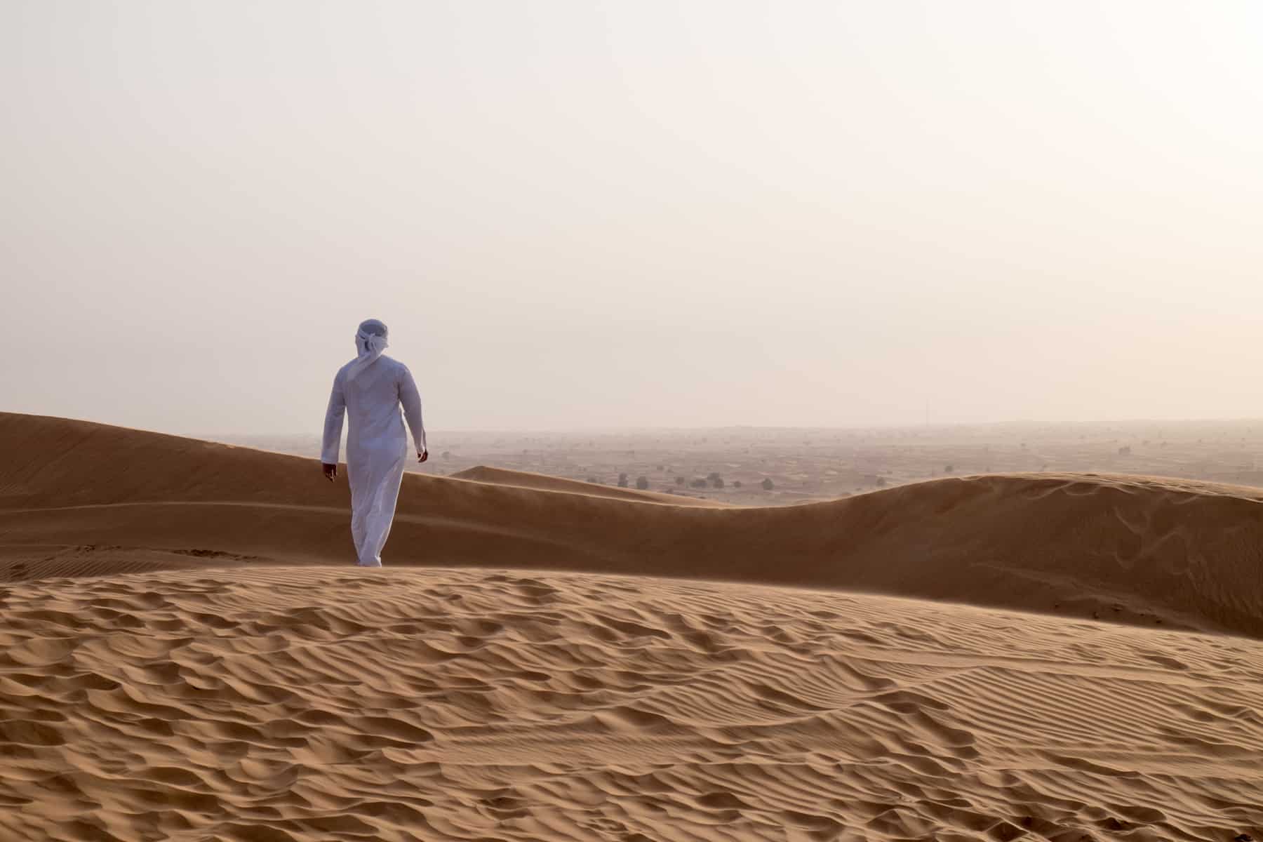 A man in a white robe walks through golden sand dunes in the Ras Al Khaimah desert at dusk.