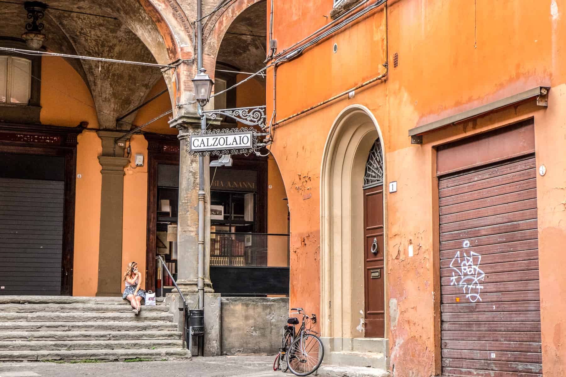 A woman on her phone sits on old stone steps in the arched corridor of an orange painted building in Bologna, Italia. 