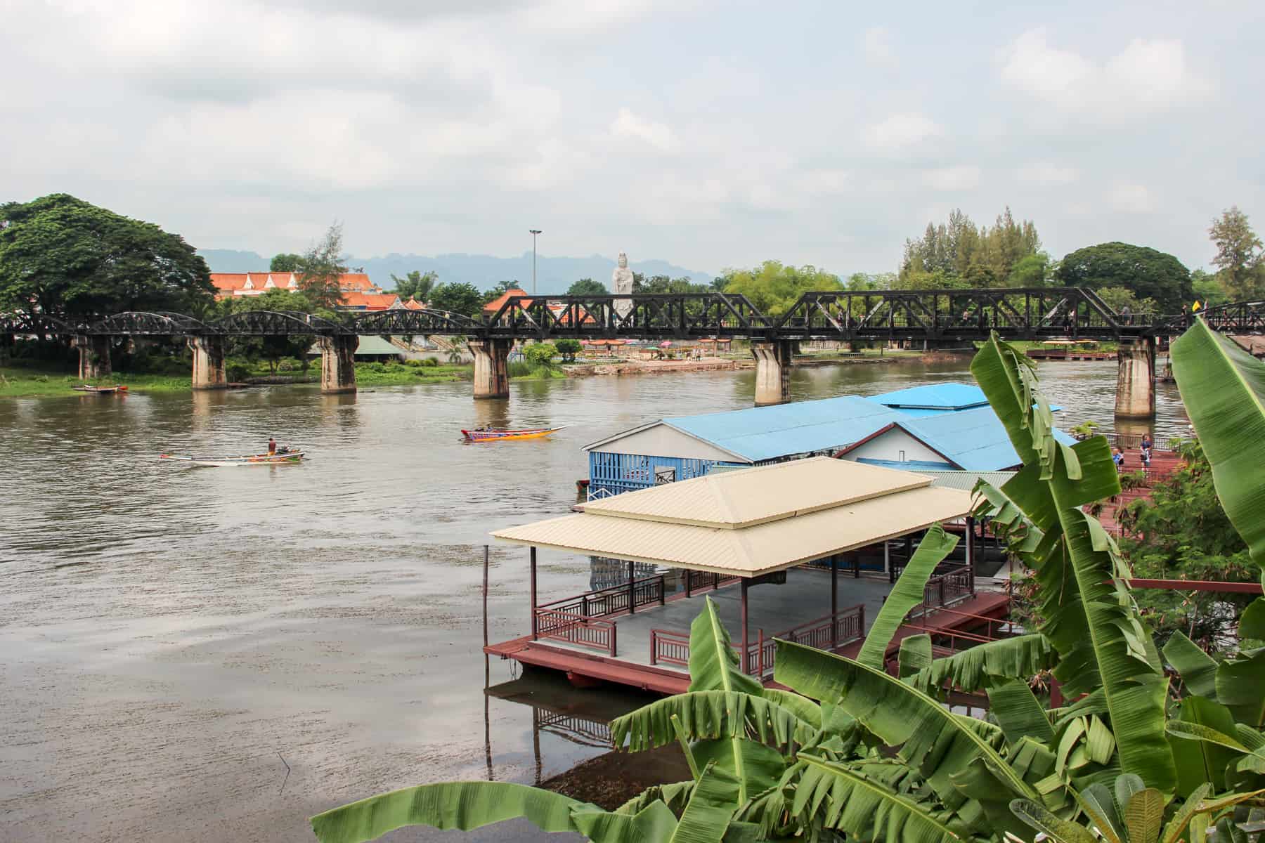 Beige and blue roof huts and two boats in front of the Bridge over the River Kwai in Thailand