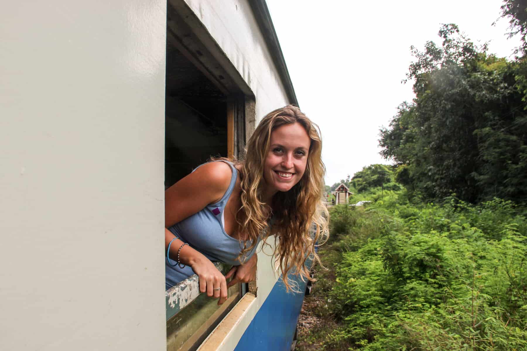 A woman leans out of a white and blue train carriage window as it journeys through rural Thai countryside