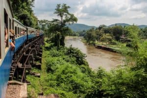 A white and blue train passes a river in a green jungle valley in Kanchanaburi, Thailand.