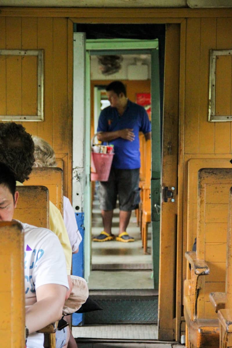 A man in a blue t-shirt and shorts, holding a pink basket on the yellow wooden interior of the Death Railway carriage.