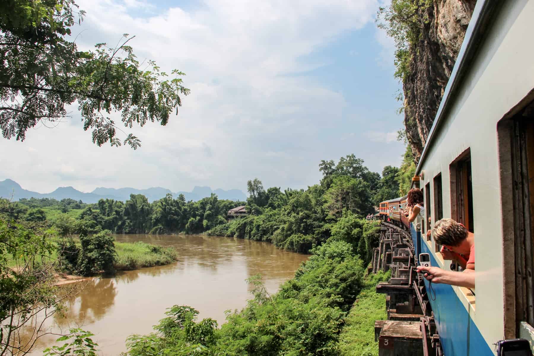 People lean out of a blue and white train - the Death Railway in Kanchanaburi – as it curves past a jungle green river bank.