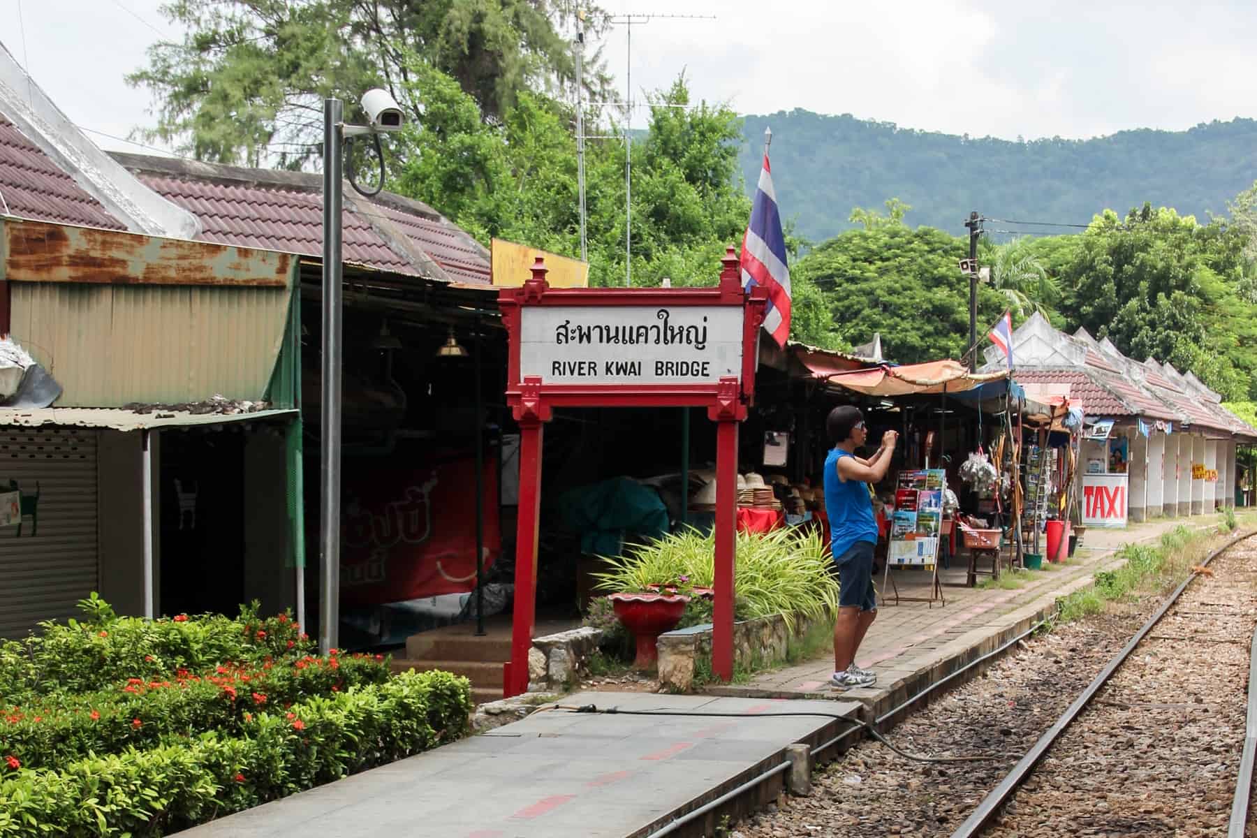 A person taking a picture on a train station platform, next to a red and white station sign that reads in black lettering "River Kwai Bridge" in Thailand