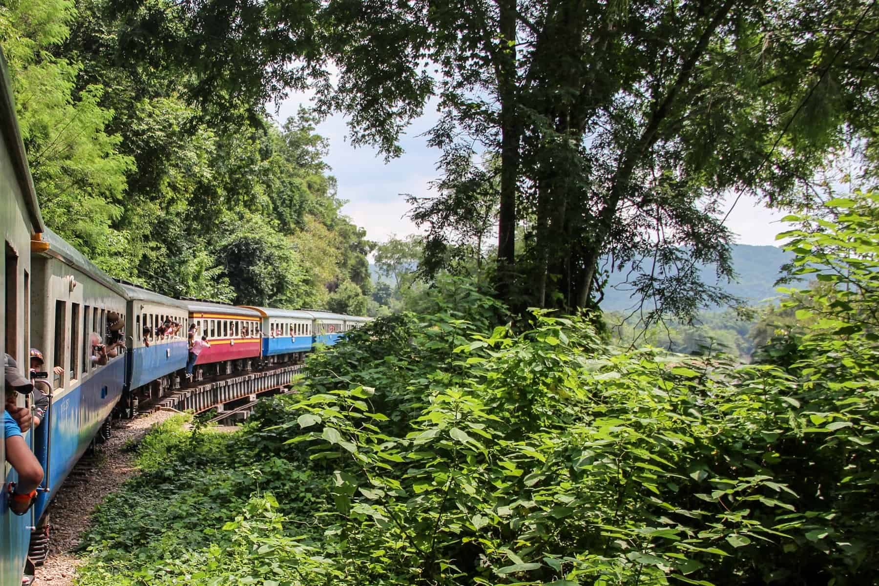 Blue, white and pink train carriages curve around a bend through the jungle green of Kanchanaburi, Thailand