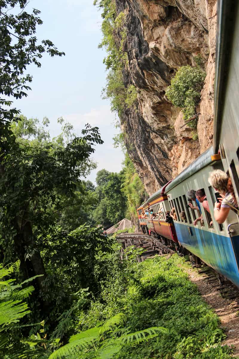 The Death Railway train in Thailand, passing golden cliffs on the right and jungle green to the left.