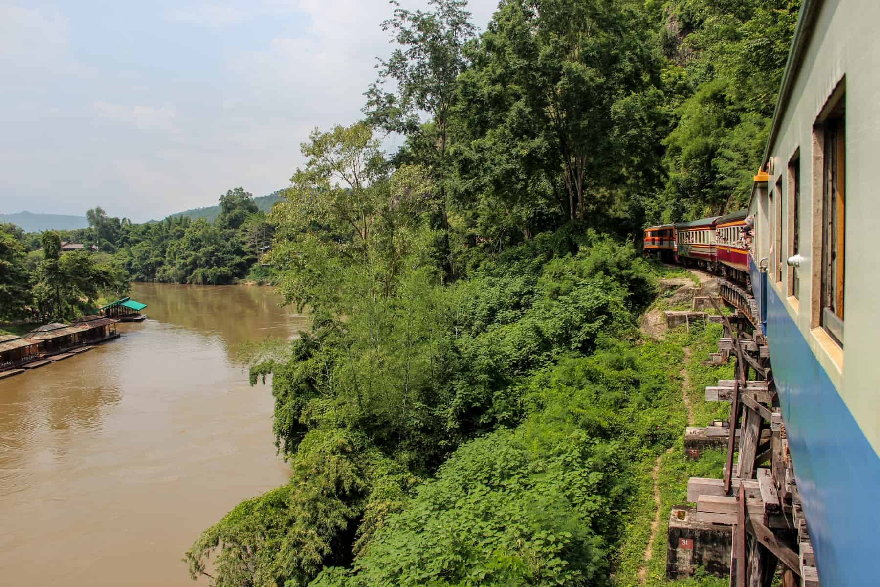 People lean out of a blue and white train - the Death Railway in Kanchanaburi – as it curves past a jungle green river bank.