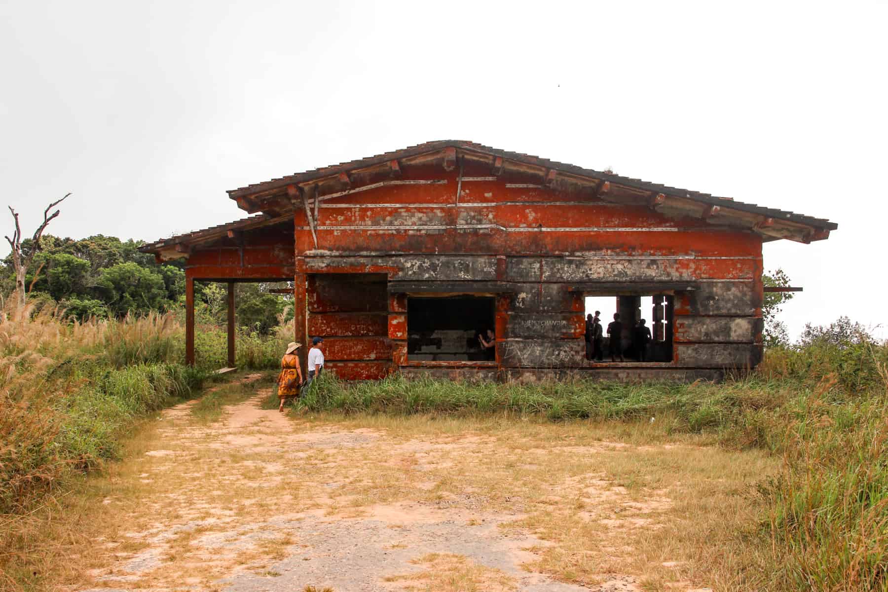 A woman in an orange dress and a man in white enter an abandoned red brick building within rugged green grassland on a hill.