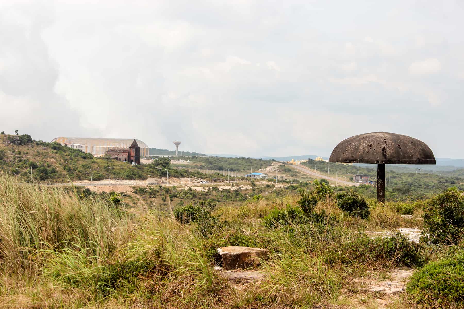 A metal mushroom shaped structure and other distant abandoned structures in unkempt nature on a hilltop in Cambodia. 