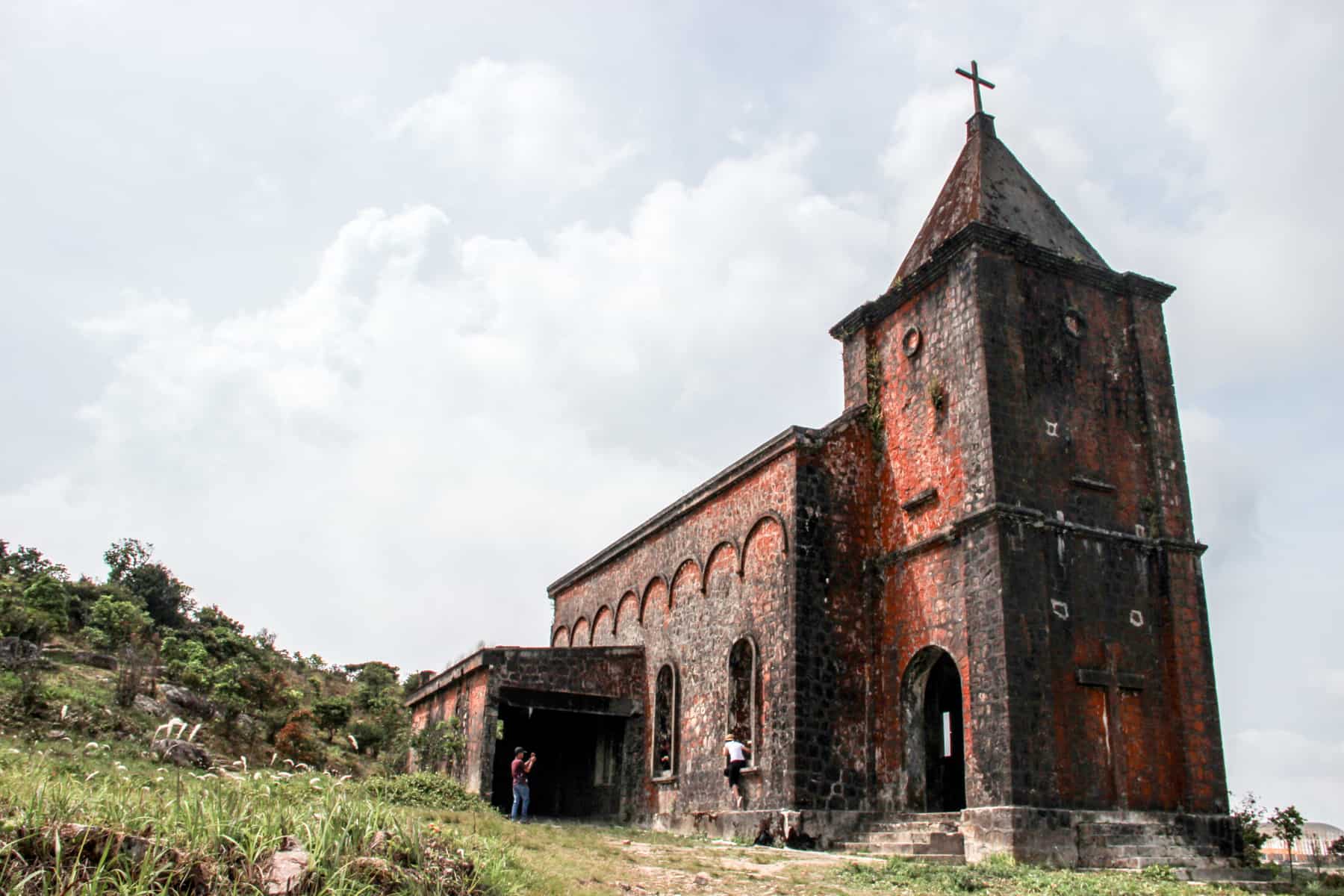 Two people outside an abandoned, blackened red stone church building on a green hilltop. 