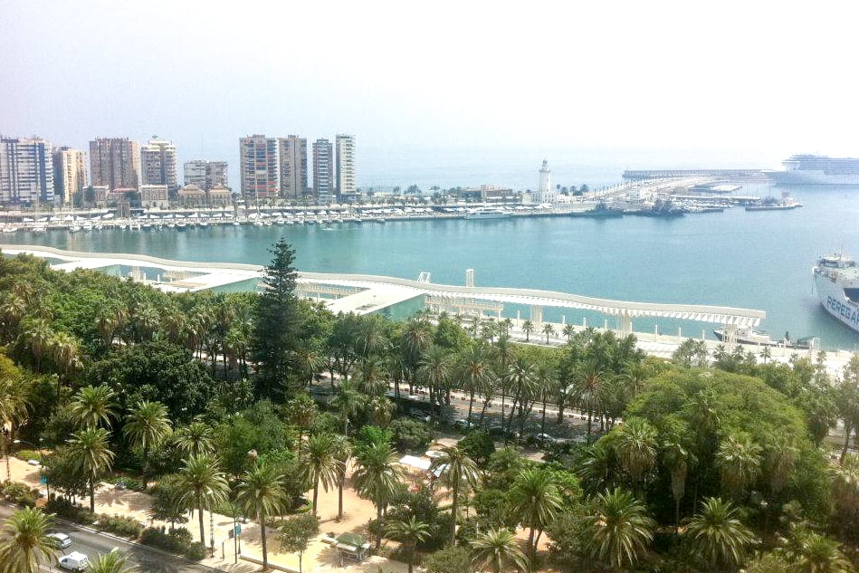 The turquoise blue waters of the coastline of Malaga, Spain next to a promenade covered by a long, white roof structure. In the background, high rise buildings can be seen on the end curve of the coastline. 