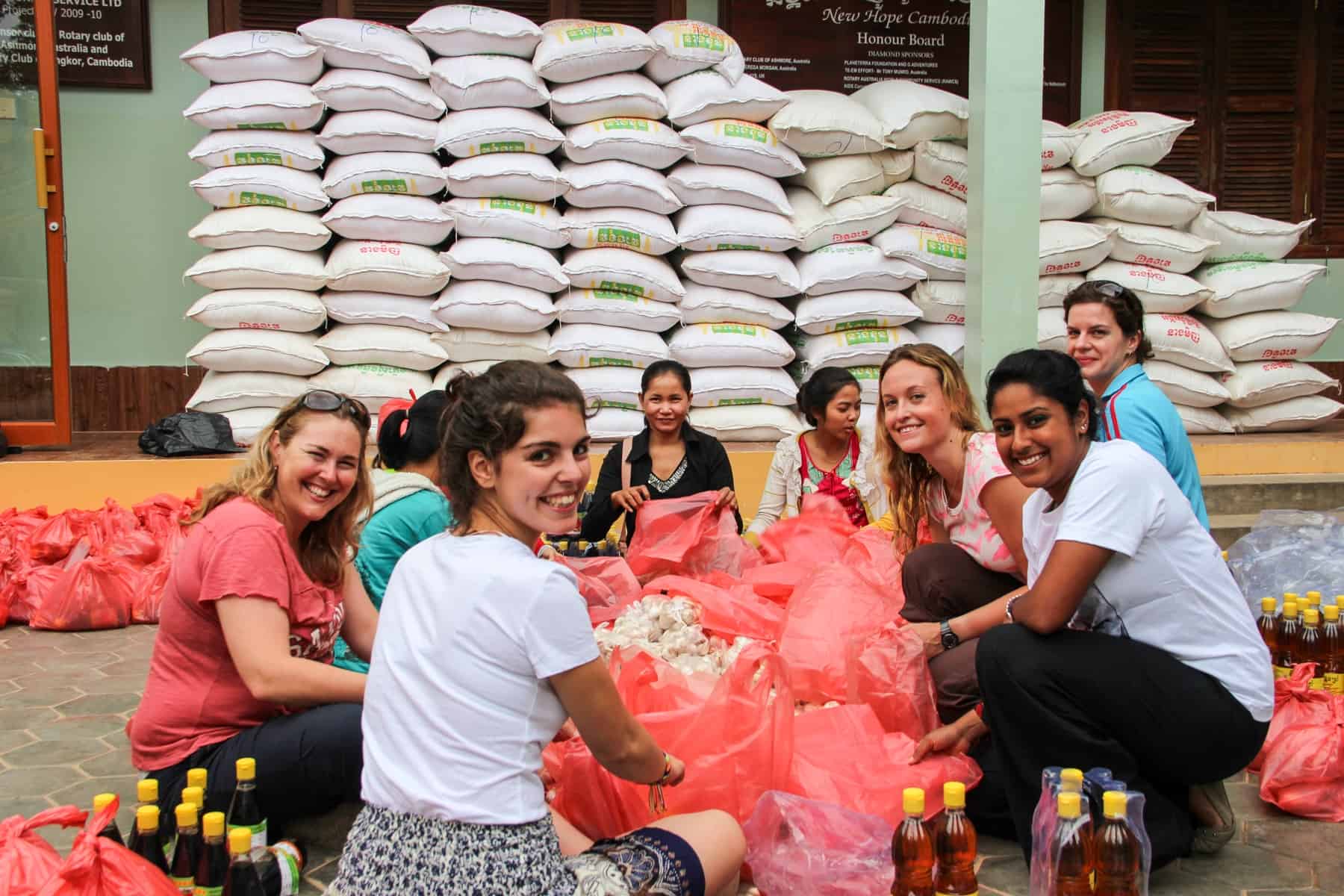 A group of women packing drinks bottles and other foods into red plastic bags. Behind them are piles of large bags of rice. 