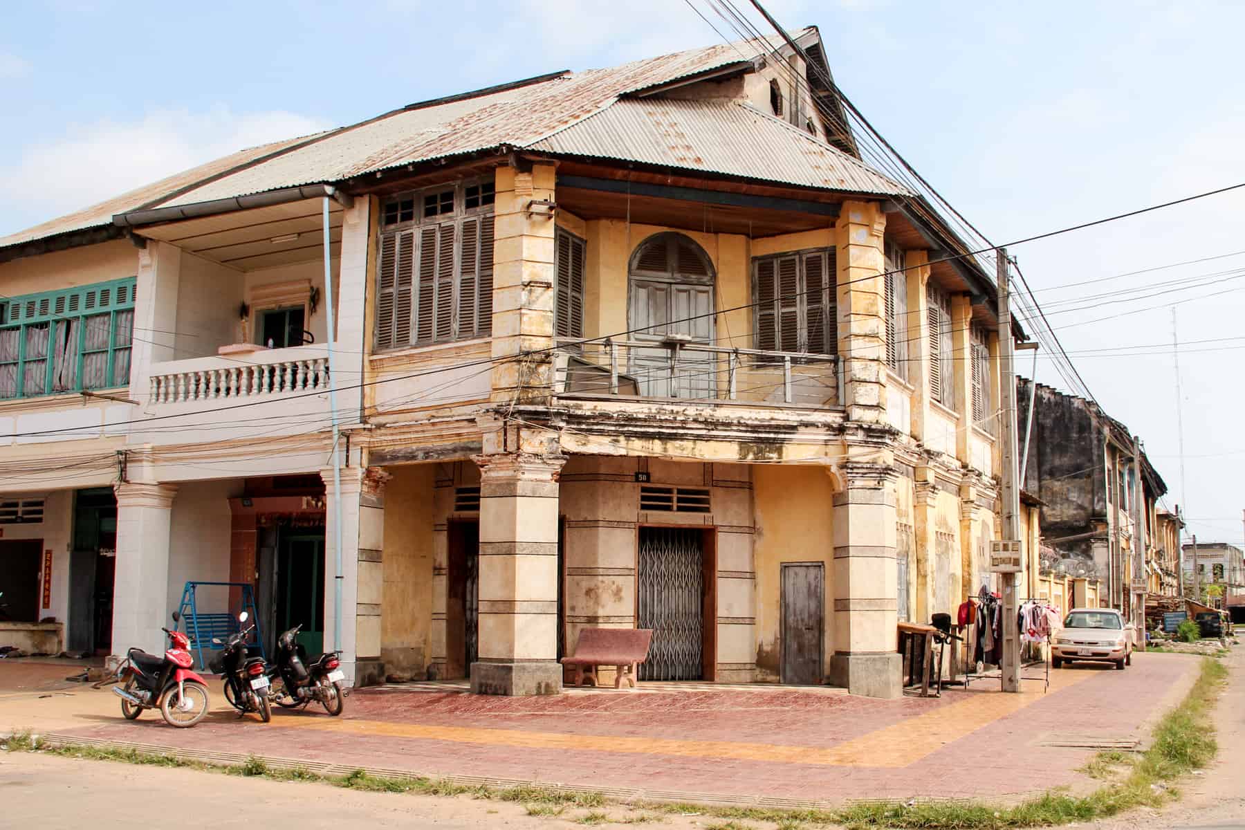 A faded yellow French colonial style mansion with wooden shutter windows, columns and a balcony on a street corner in Kampot city.