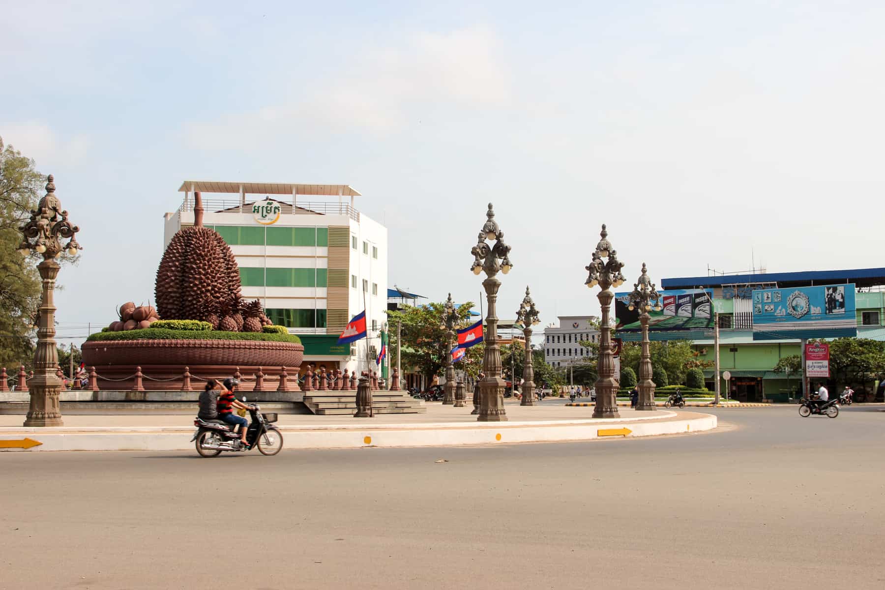 Two motorbikes circle a huge roundabout in Kampot in Cambodia which has in its centre a huge Durian fruit artwork. Behind the roundabout are modern block buildings. 