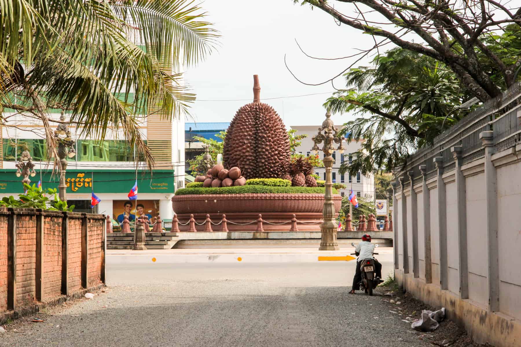 View from a side street where a man on a motorbike looks towards a roundabout with a huge carved Durian fruit sculpture.