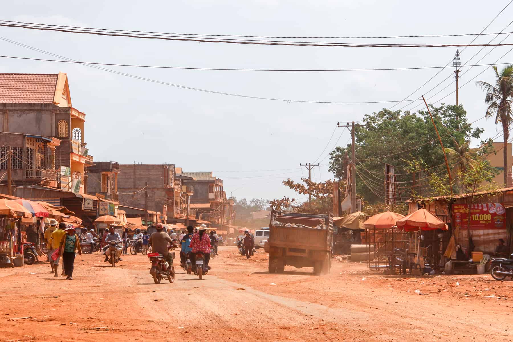 People, motorbikes, trucks and street stalls fill a wide street covered in ochre orange dusty sand. As are the buildings that line the street. 