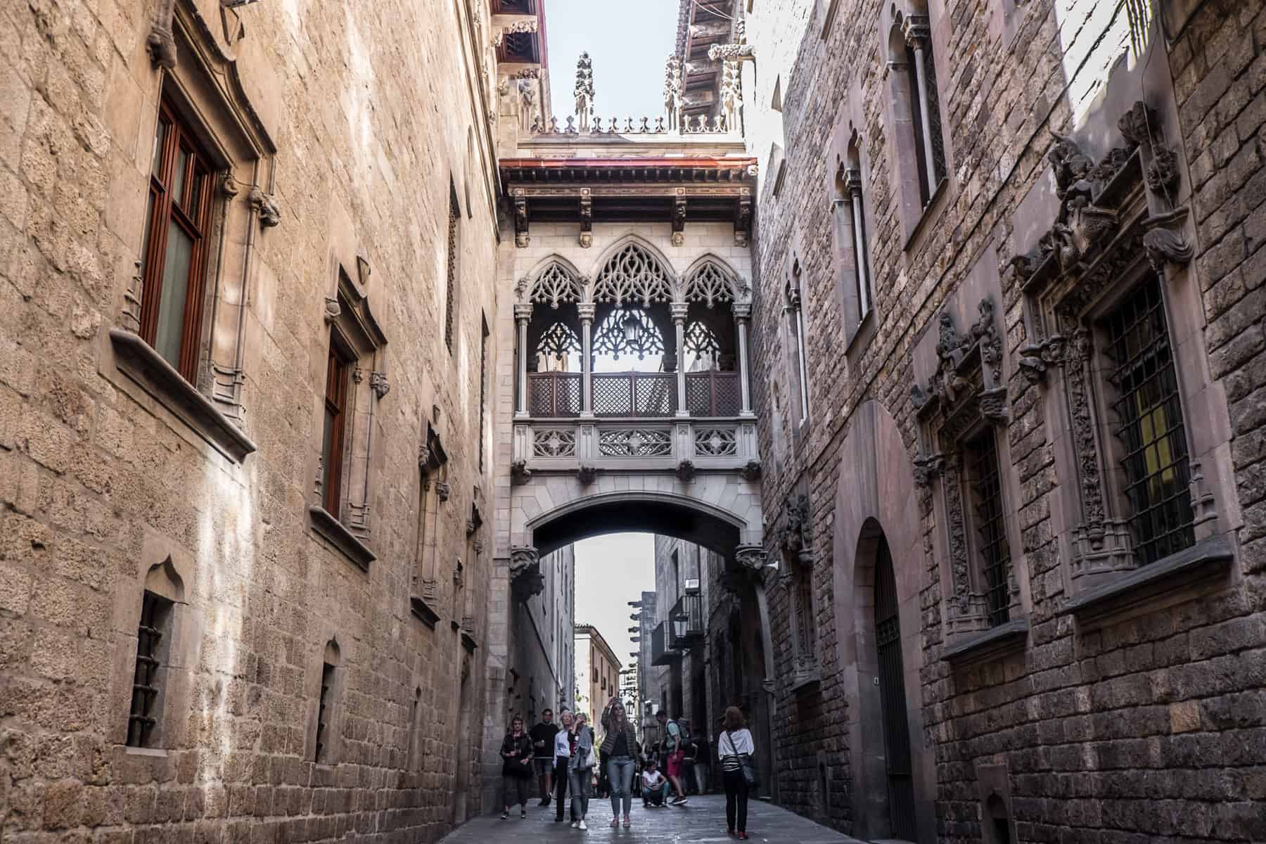 A narrow, delicately carved balcony connects two old golden and brown stone buildings on a long street in Barcelona. People are walking underneath the structure. 