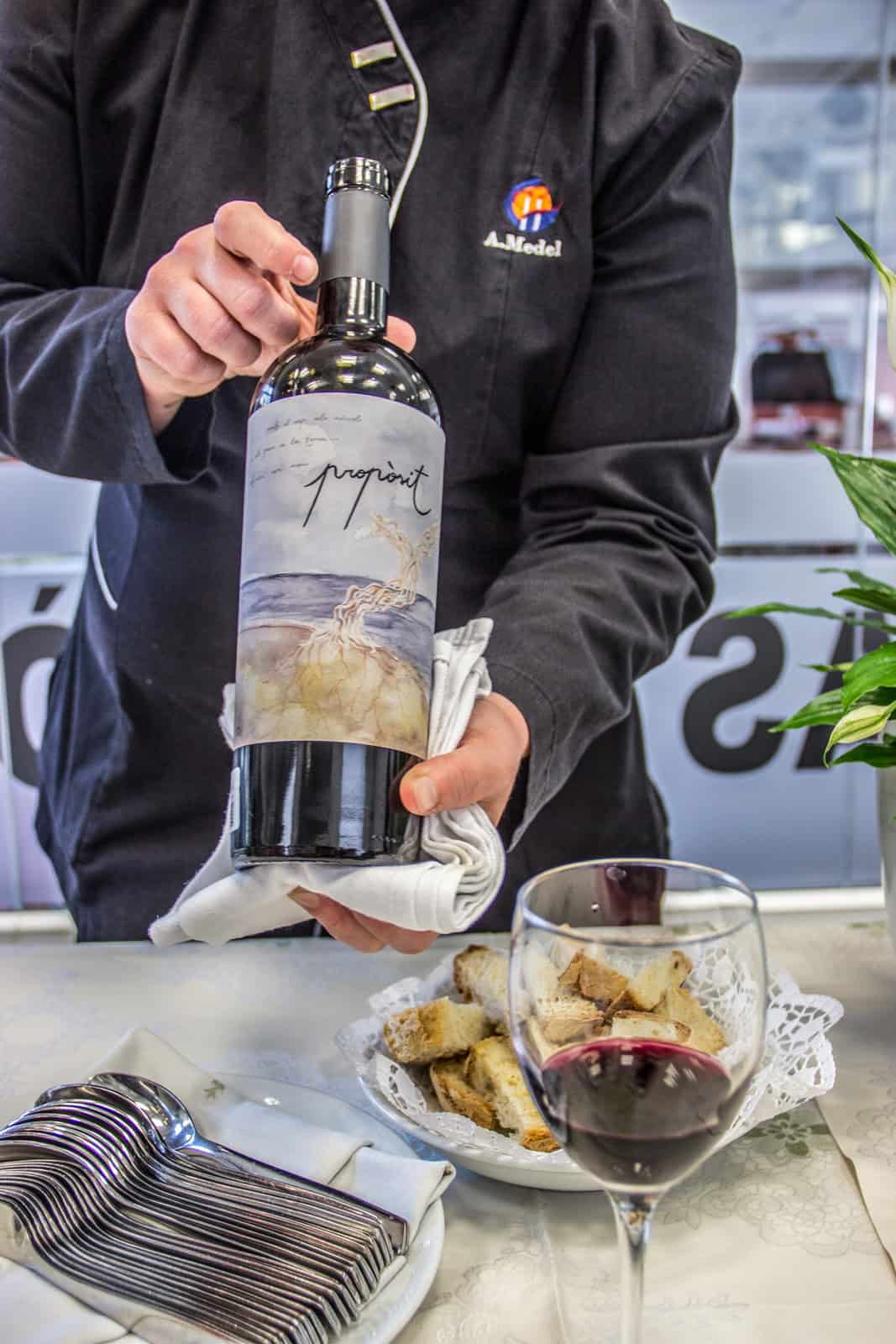 A waiter holds a bottle of wine; on the table is a glass of red wine and some a small pile of bite-size bread pieces. 