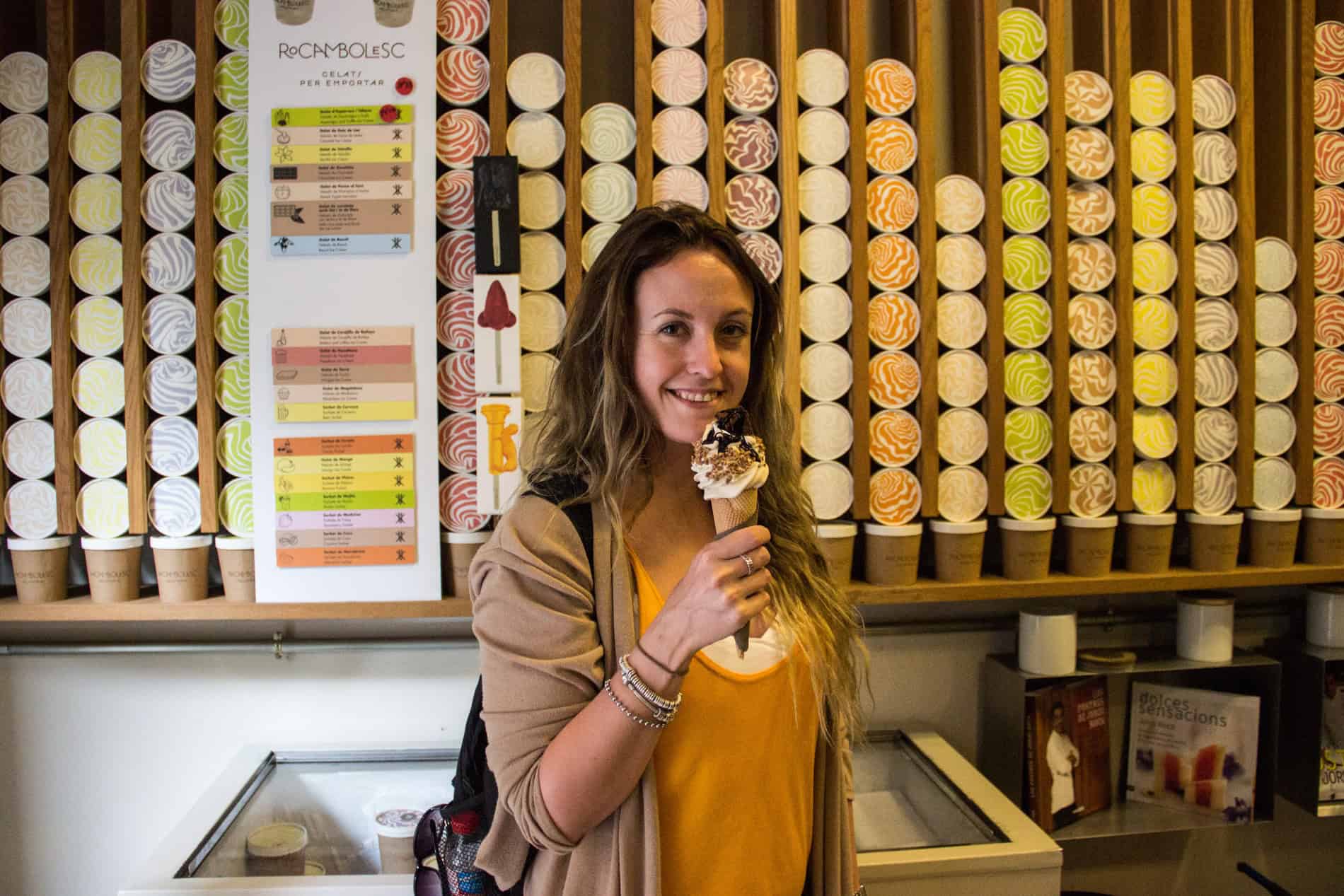 A woman with long hair holds an ice cream cone while sanding in front of a wall of colourful ice cream tubs. 