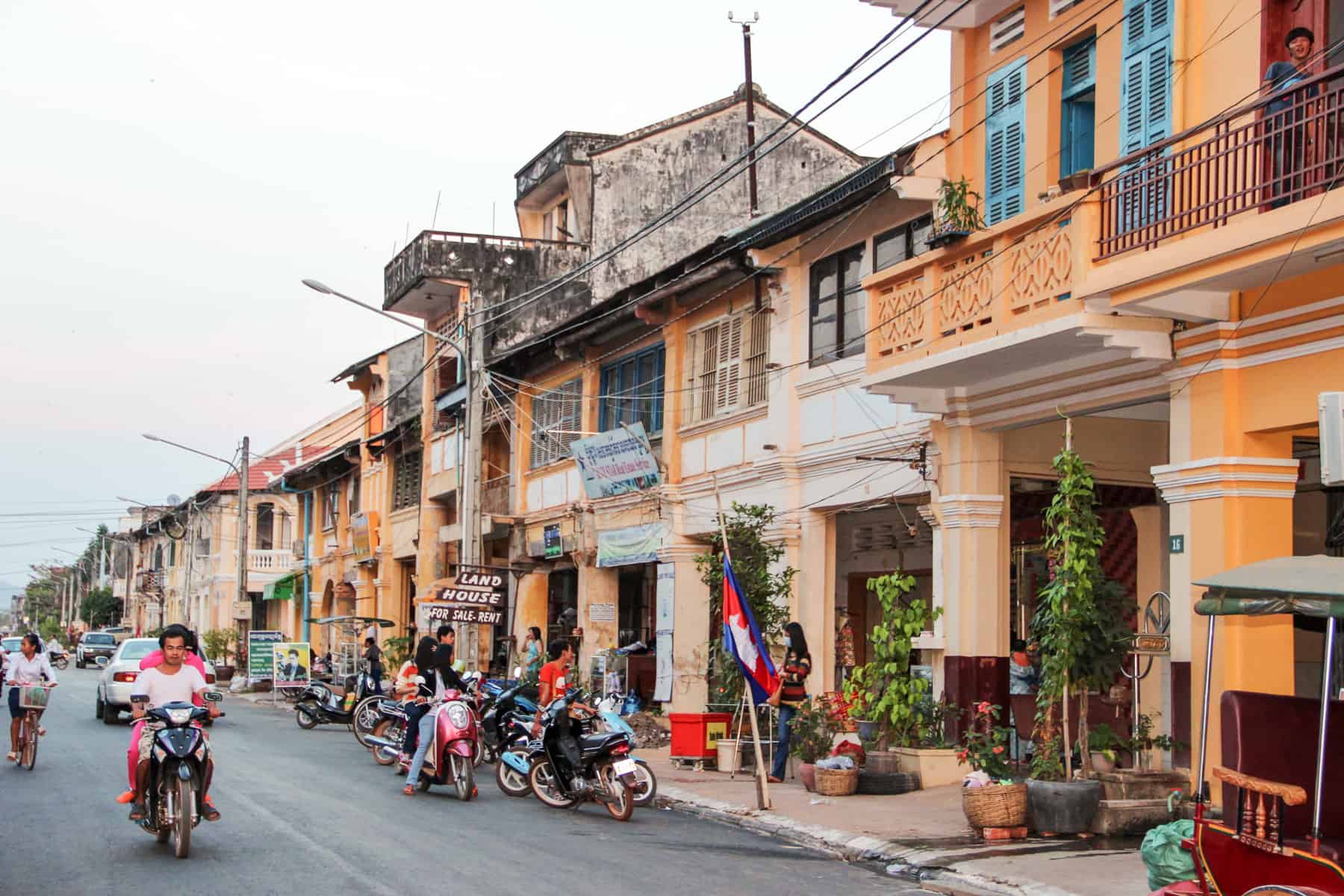 People on motorbikes ride past a row of orange painted houses in French colonial architectural style in Kampot in Cambodia.