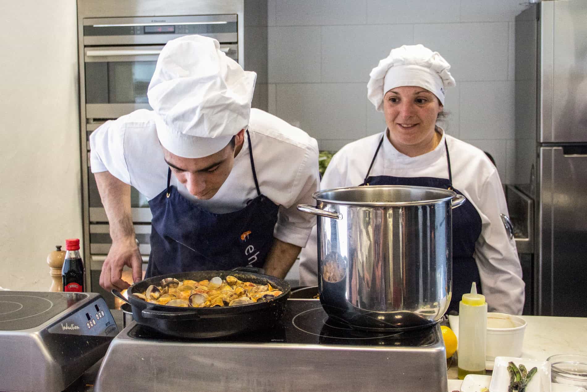 A man and woman in chefs whites stand behind a large pot and a sizzling pan on a stove. 