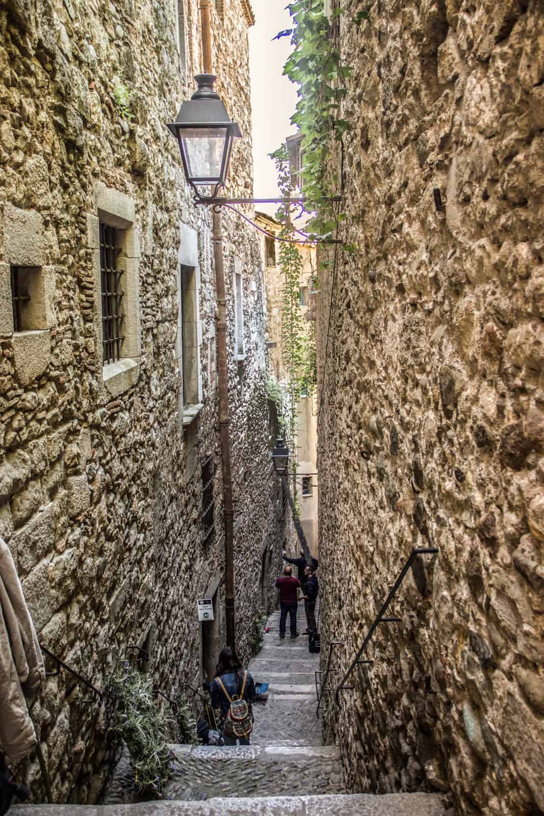 People walking down long steps in a very narrow street between old rounded-stone buildings and walls. 