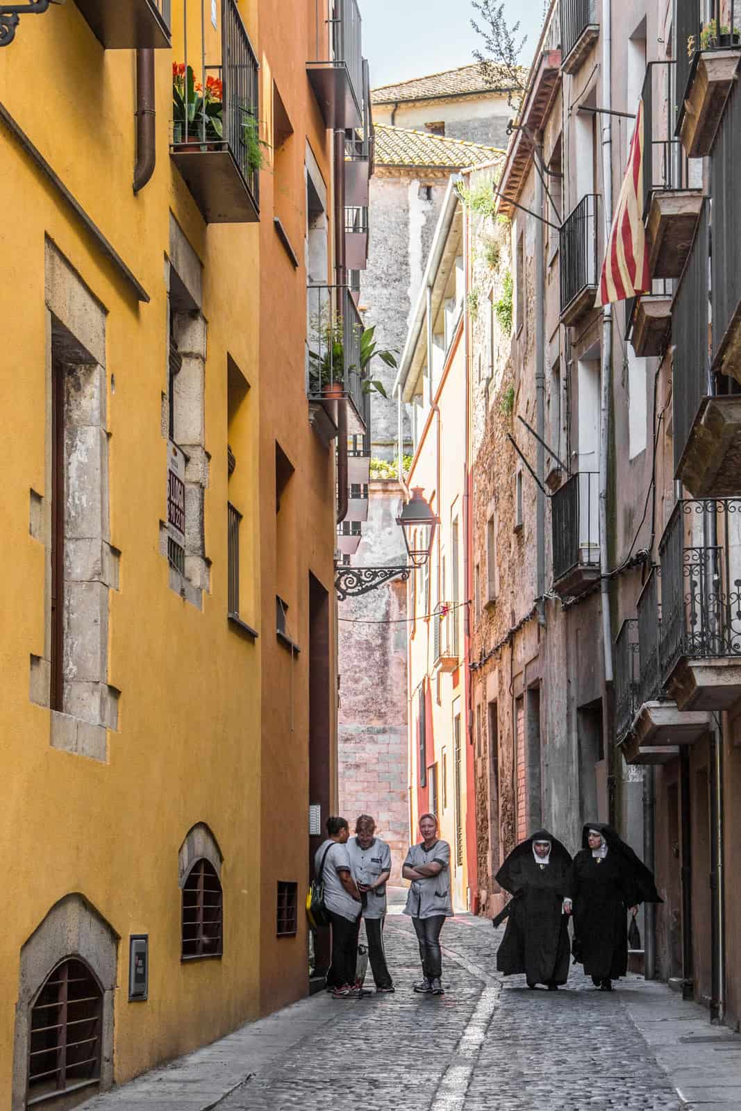 Women is white shorts and a group of nuns in a narrow cobblestoned street in Girona with yellow buildings and old stone structures. 