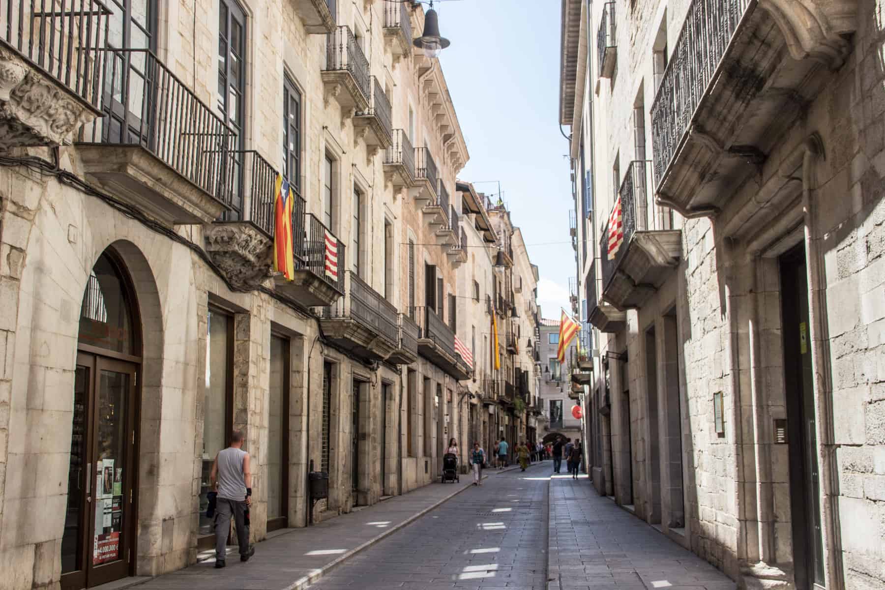 A paved street lined either side with creamy white stoned buildings. Some of the balconies have the Catalonia flag hanging from them, 