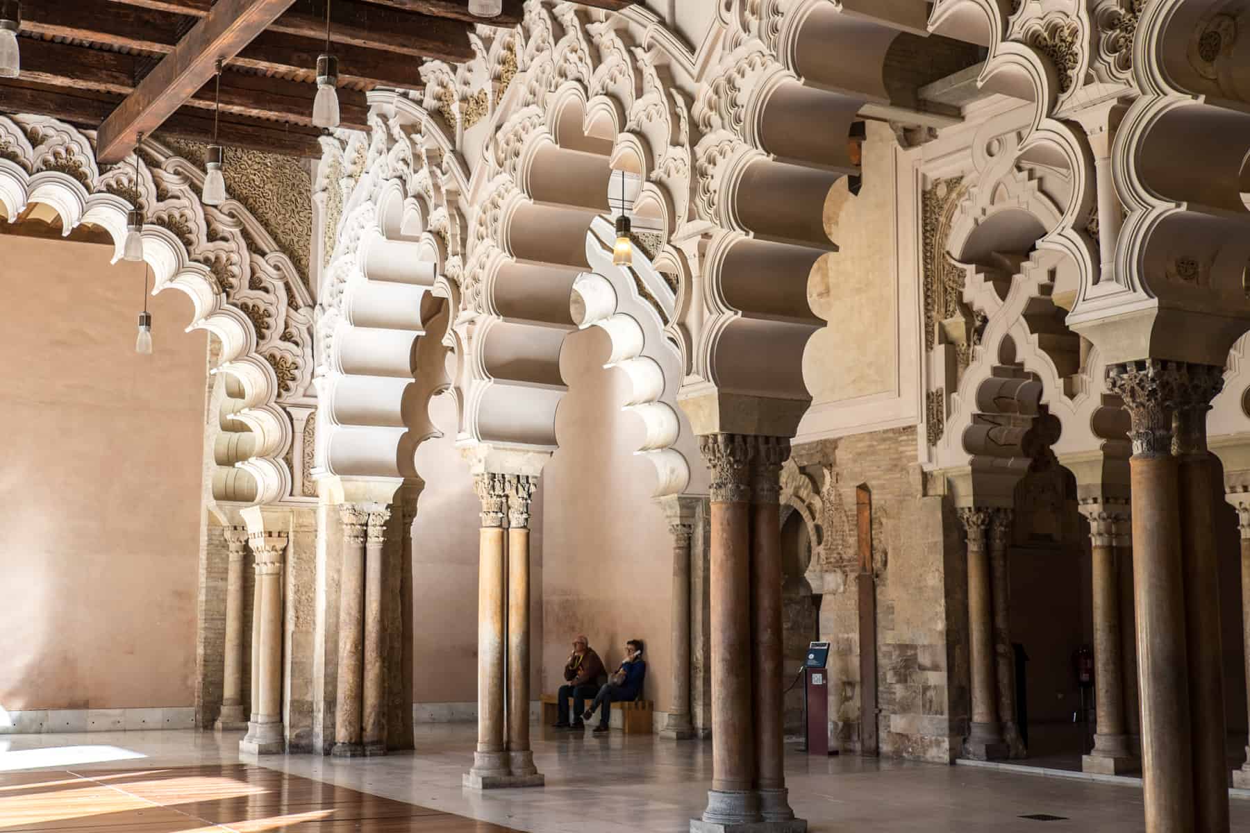Two people sitting behind a row of carved, creamy white Mudéjar art archways found in an Islamic palace in Zaragoza, Spain. 