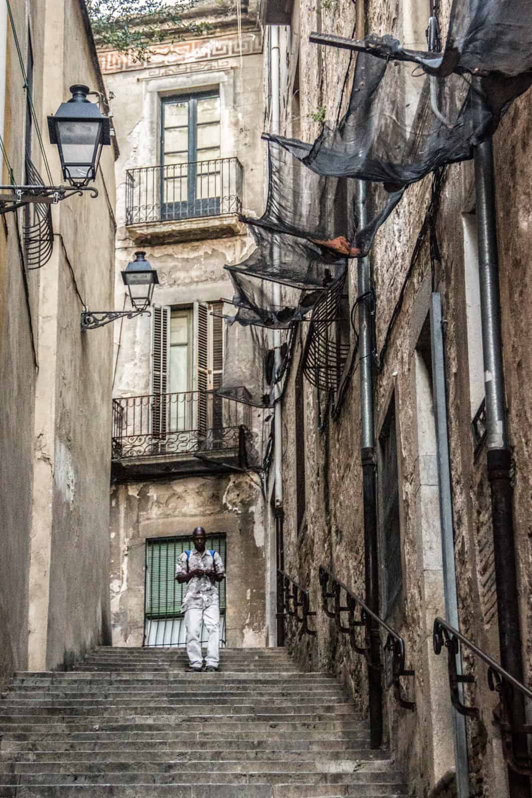 A man in white walks down stone stairs on a narrow timeworn street with black lamps and black protective netting over the windows. 