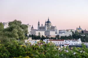 A silvery white palace complex with dark grey dome roof and spires is elevated above square city buildings in Madrid. The view is from within the foliage of parkland, and at a distance.