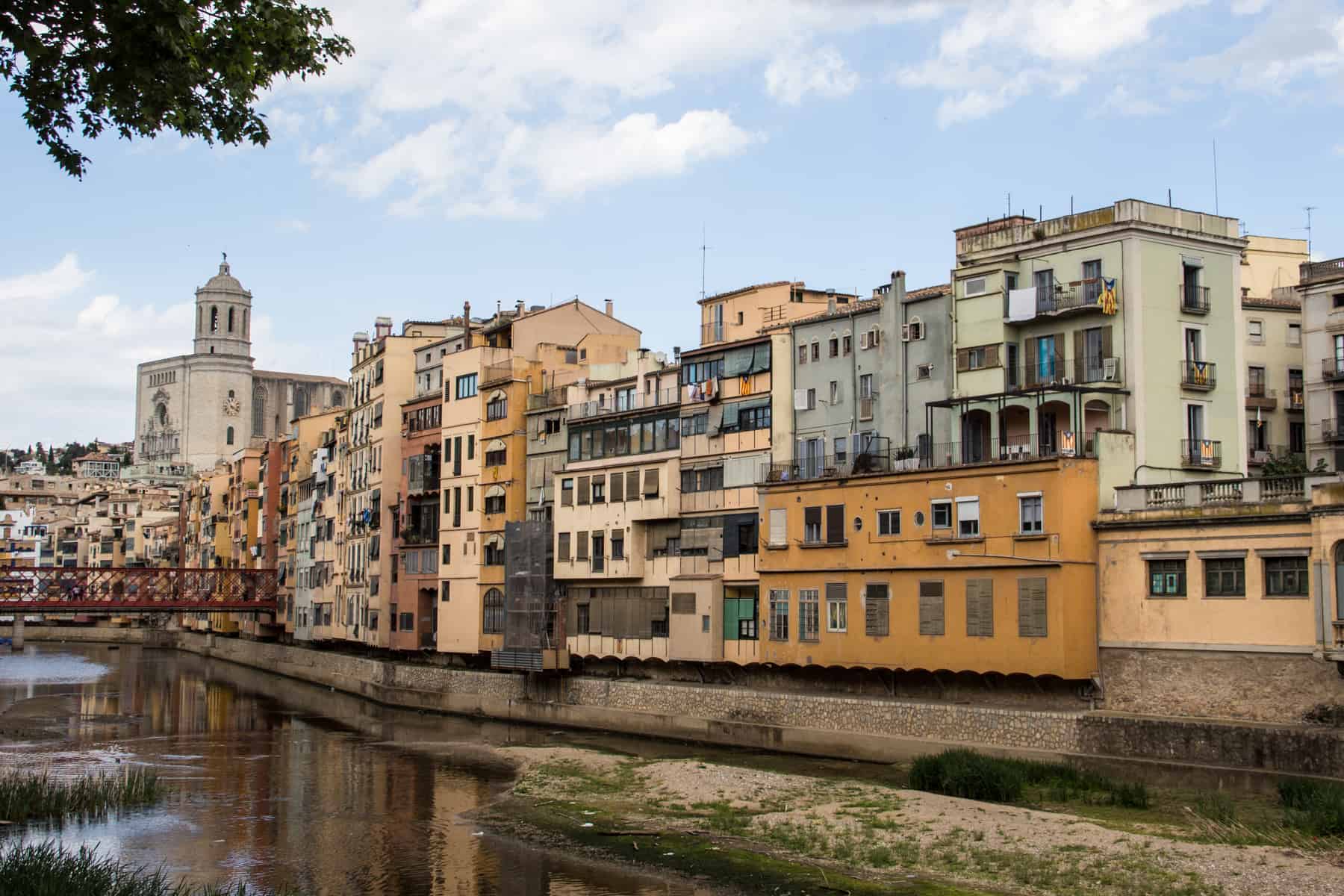 A row of square and rectangular buildings in shades of yellow, orange an mint sit next to a river. At the far end is a small red iron bridge, and beyond that the white spire of a church. 