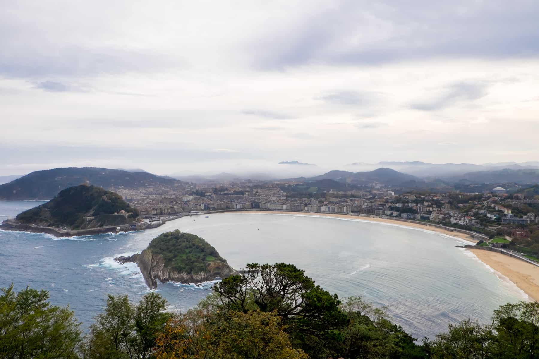 The semi circular coastline of San Sebastian in Northern Spain. To the left are two island mounds, covered in green, and to the right, yellow sand beaches. The city buildings are in the middle. 