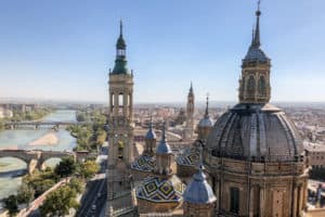 Blue and yellow mosaic domed rooftops in front of a wider golden building city view of Zaragoza, next to a turquoise river. An elevated view from the rooftop of a cathedral.