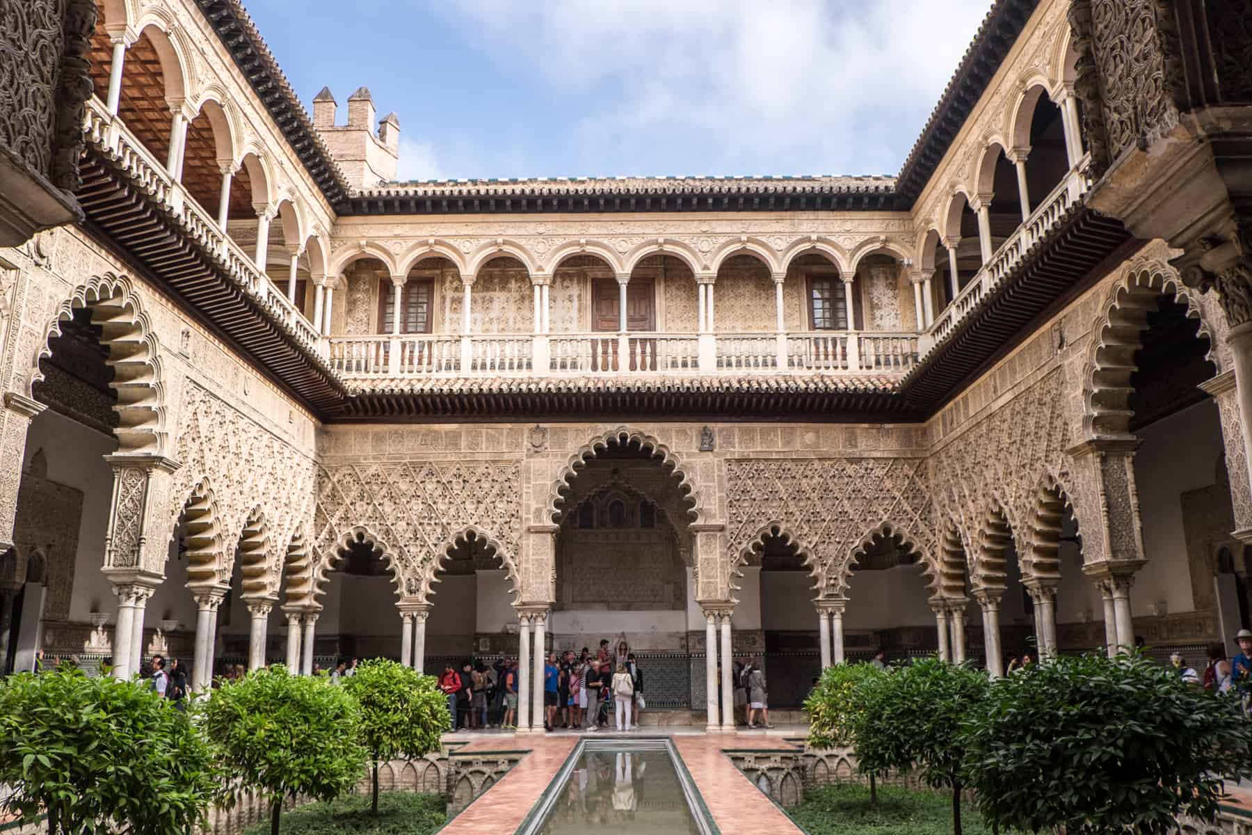 The two tiered intricately carved and curved archways and balconies in the Mudéjar architectural palace of the Real Alcázar of Seville. In this open courtyard a pool of water cut through its middle, and people can be found standing in the corridors of the lower floor.. 