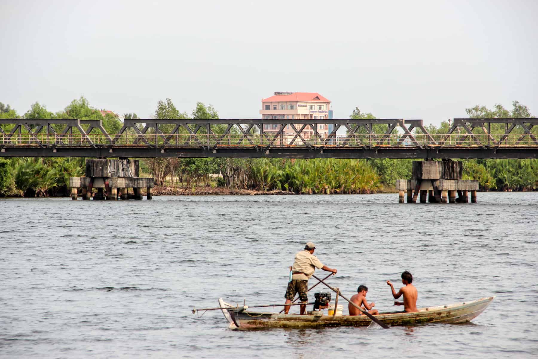 Three men on a wooden boat in a wide river in front of an old bridge. Behind is a solitary white block house with an orange roof, that sits within jungle green.