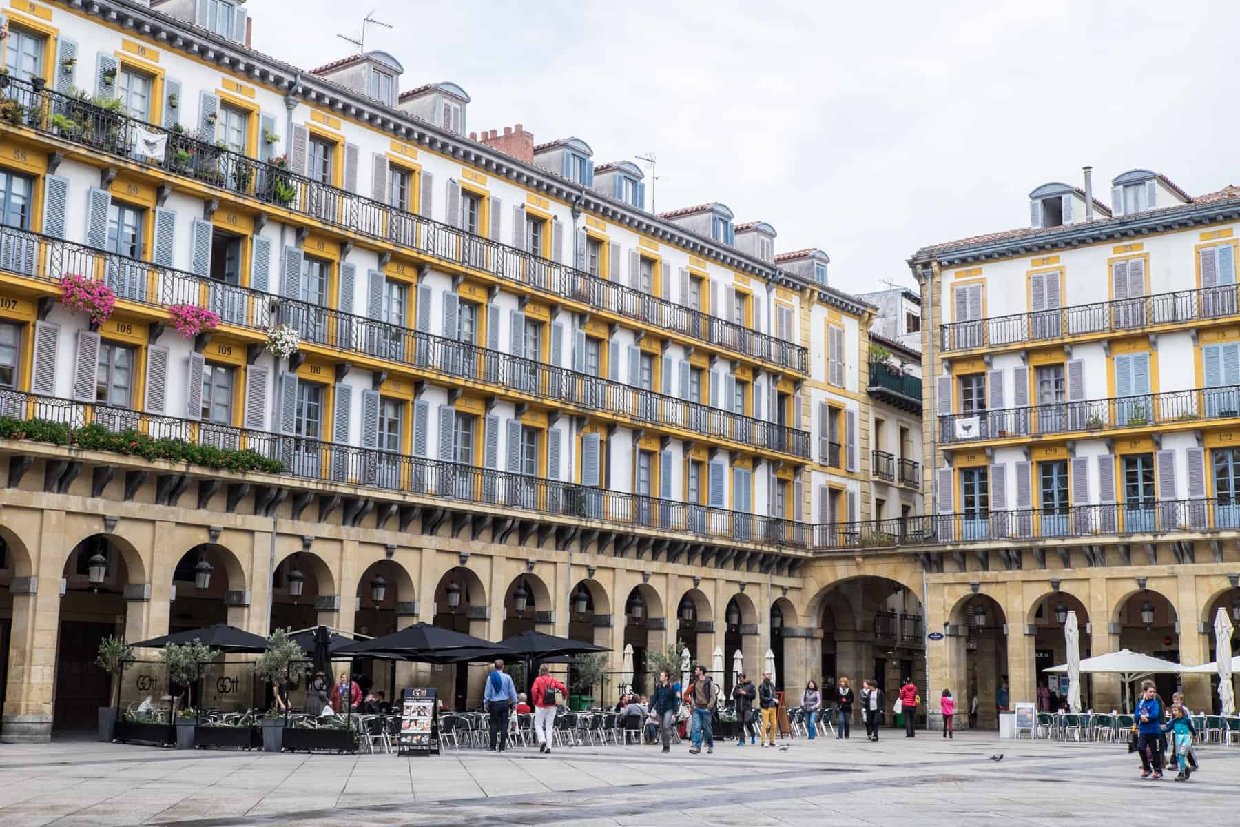 People walking through a public square whose surrounding building consists of rows of arched porticos on the lower floor and three rows of yellow and grey windows. 