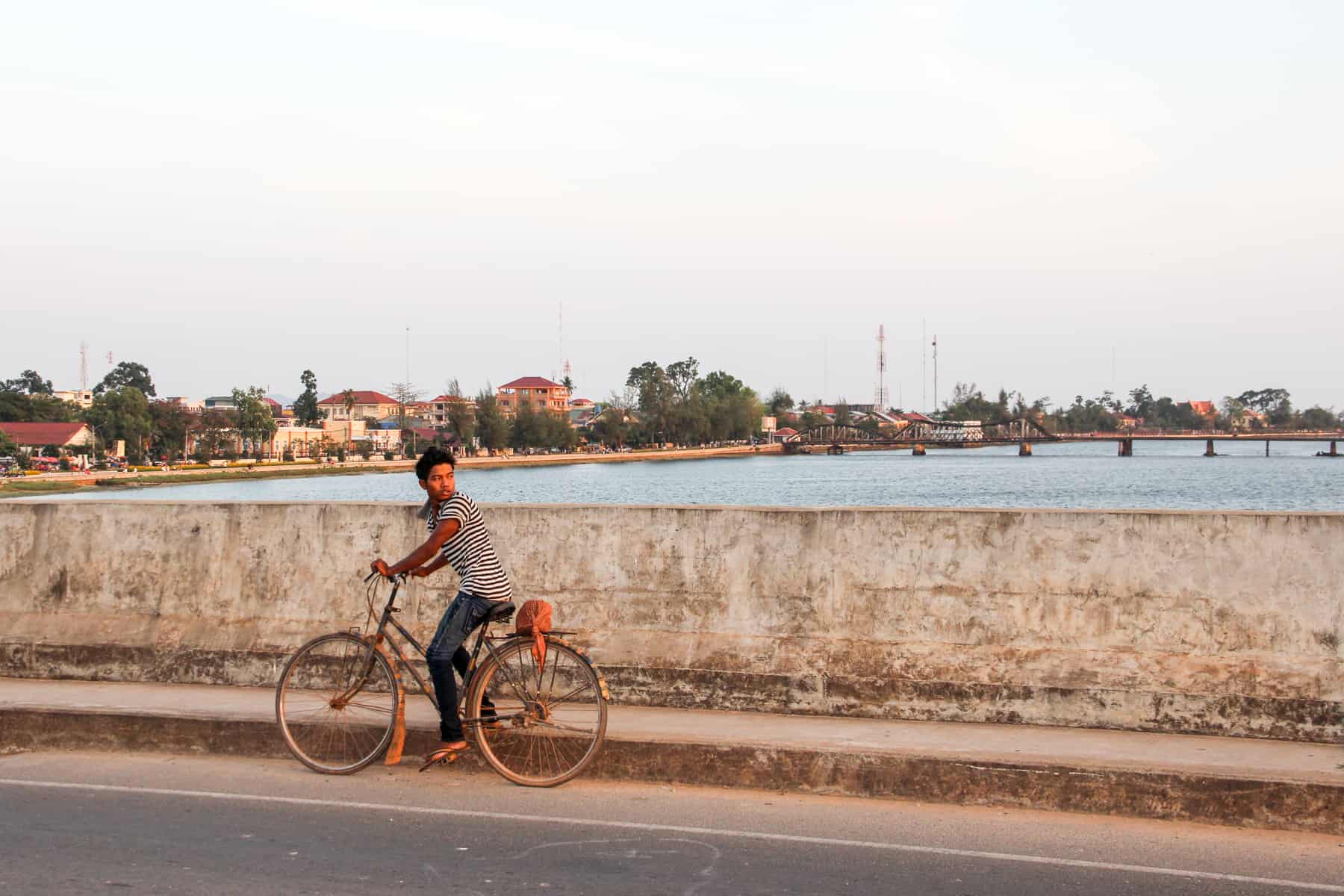 A boy in a striped t-shirt rides a bicycle on a beige stone wall bridge that crosses a light blue river. In the background is another bridge and orange buildings lining the riverside. 