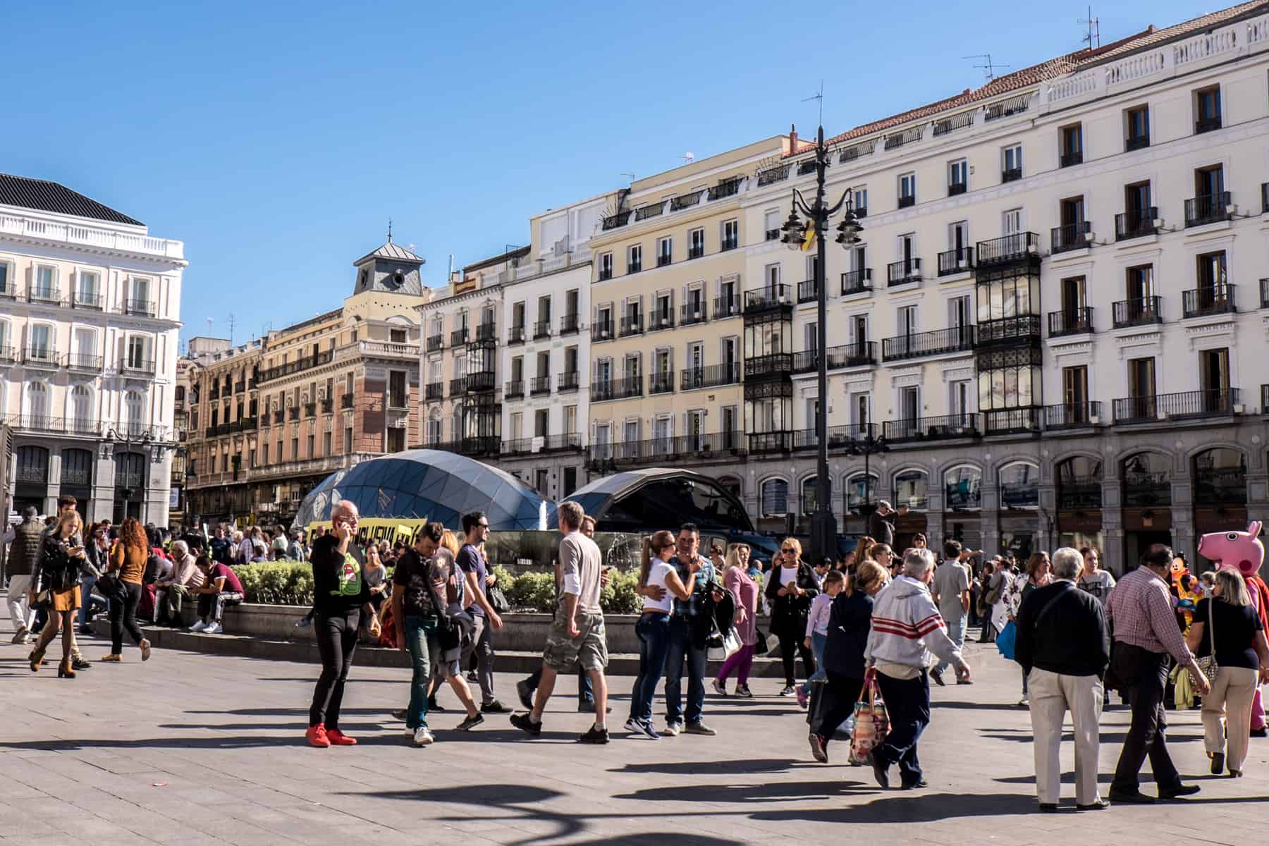 People dispersed in the wide pathways of a public square in Madrid. In the middle of the square is a large oval flowerbed and behind a rows of rectangular, multi-windowed buildings in a blend of white and cream hues.