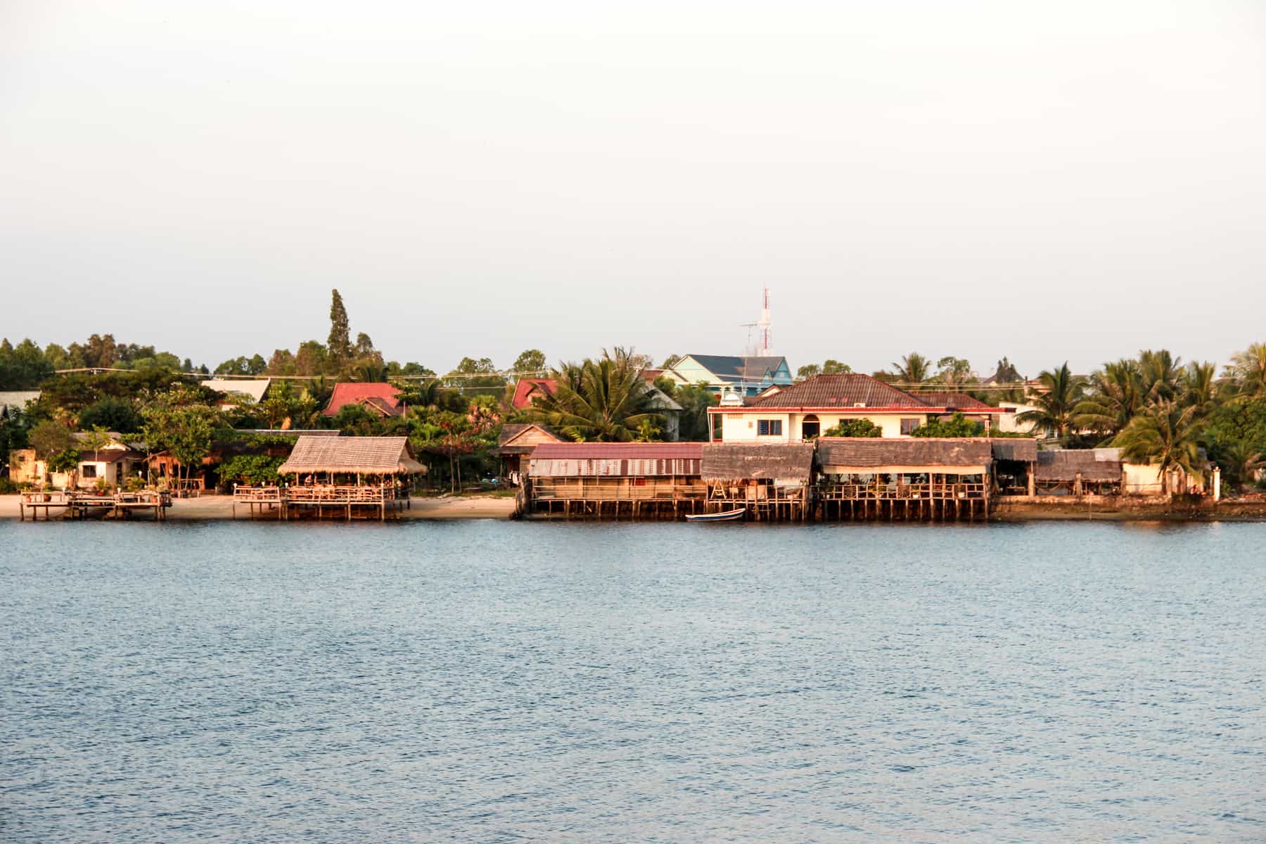 Wooden huts on stilts on the riverside of Kampot in Cambodia. Behind them are white houses with orange roofs set within green forest. 