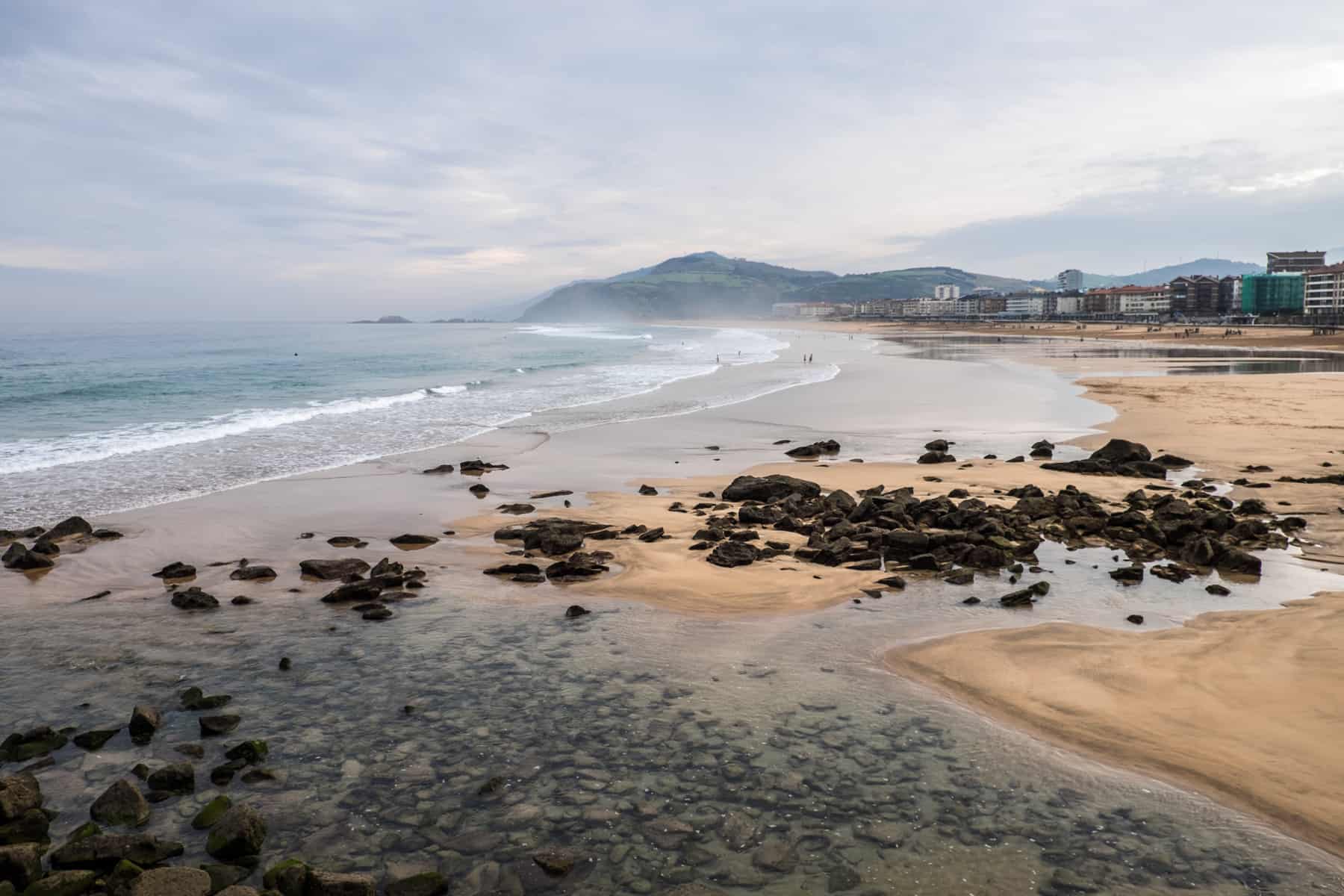 Large rocks strewn on a yellow sand beach of La Concha in San Sebastian. Modern city buildings and rolling hills can be seen in the background. 