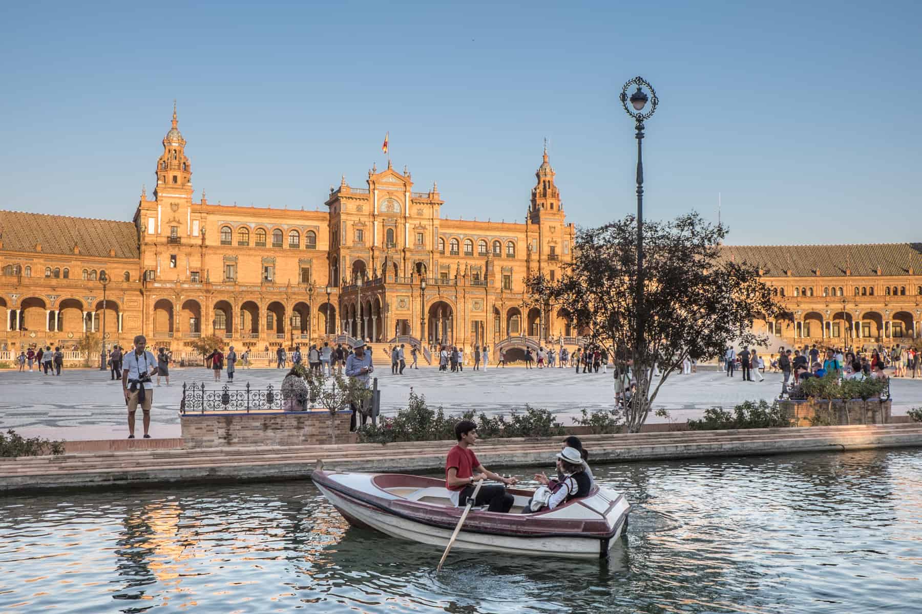 A man rows a boat with two passengers on a small manmade river in front of a colossal golden building that glows in the sun. People are walking in the wide plaza in front of the building and a man stands next to the river. 