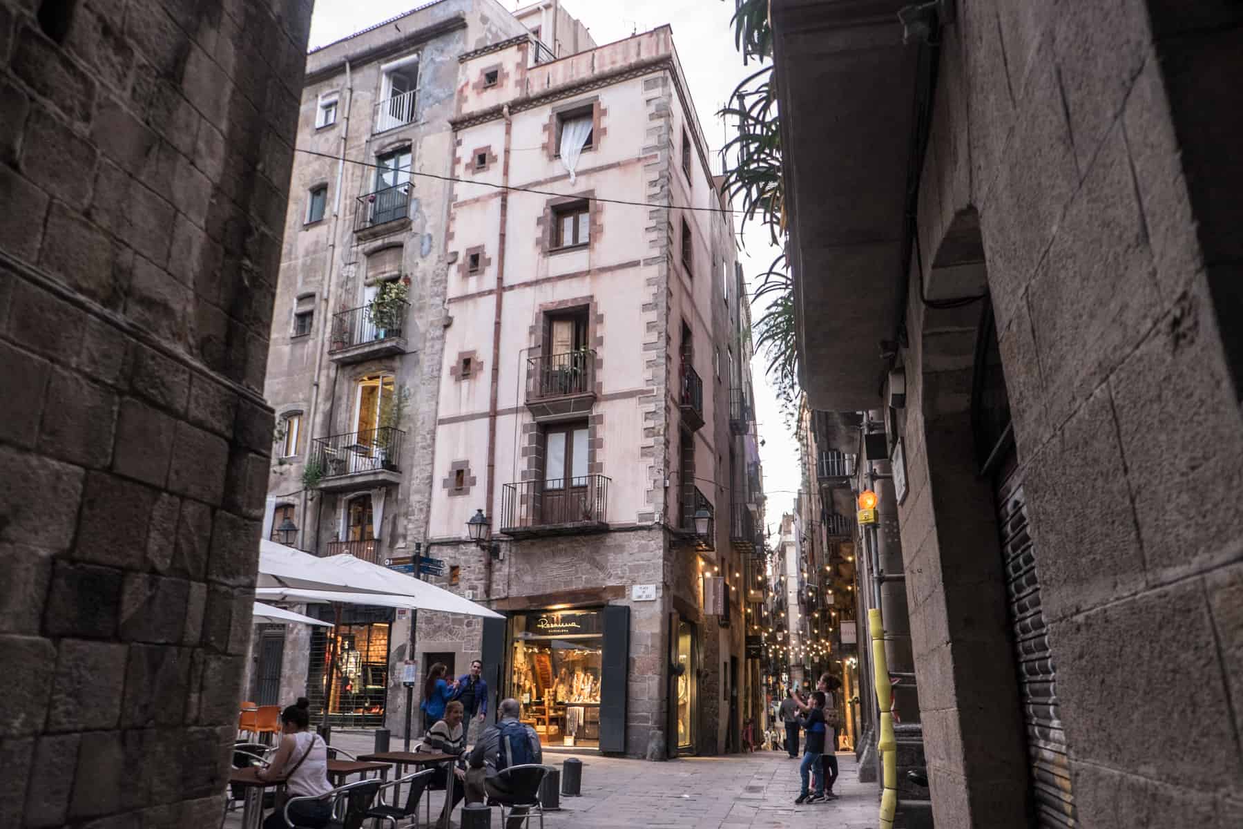 View from a street, looking into a small square that connects to other alleyways in Barcelona. People are sitting in the square at tables under umbrellas. 