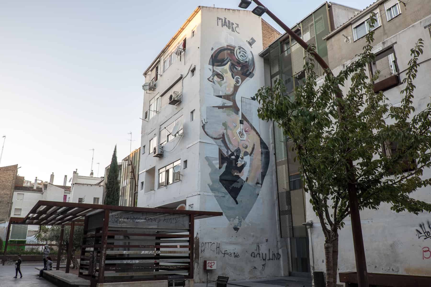 A neighbourhood of apartment buildings, once of which features a large work of street art on the side of its facade. In front is a brown wooden seating area and a tree. 